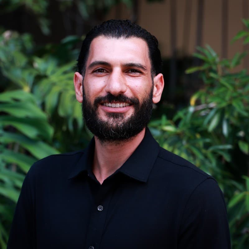 Smiling man with dark beard and black shirt standing before green foliage.